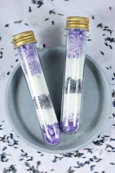 Two glass tubes of layered purple and white bath salts with gold lids, on a bed of dried lavender.