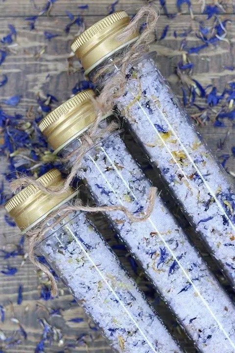 Three glass tubes of bath salts with gold lids and twine, among dried blue flowers.